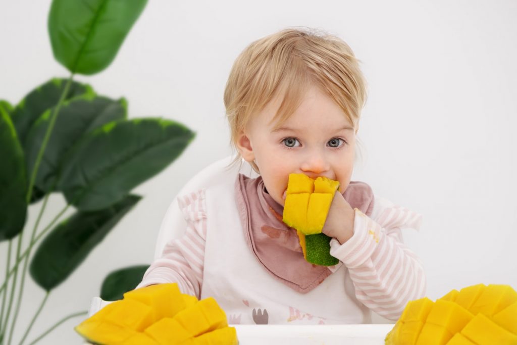 blonde baby eating slices of mango green leaves on side